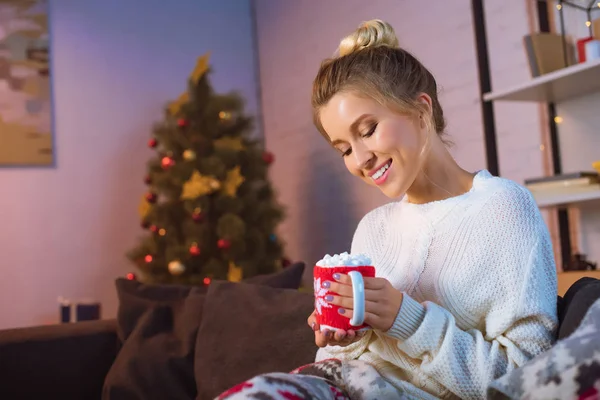 Smiling young blonde woman covered in blanket sitting on couch and holding cup of hot cocoa with marshmallows at christmas time — Stock Photo