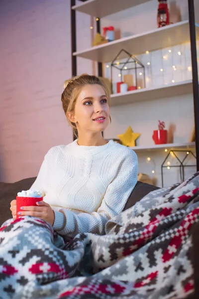 Jeune femme blonde couverte de couverture assise sur le canapé et tenant une tasse de cacao chaud avec des guimauves à Noël — Photo de stock