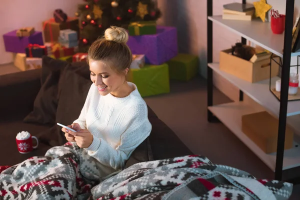 Усміхнена молода блондинка сидить на дивані і використовує смартфон у різдвяний час — стокове фото