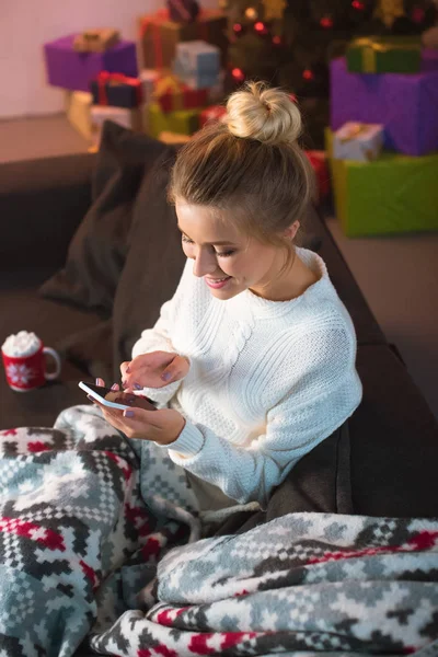 Lächelnde junge blonde Frau, die zur Weihnachtszeit auf der Couch sitzt und ihr Smartphone benutzt — Stockfoto