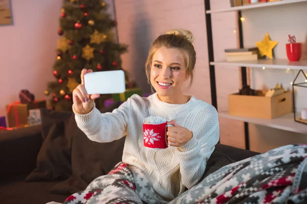 Молодая счастливая блондинка, покрытая одеялом, сидит на диване, держит чашку горячего какао с зефиром и делает селфи на смартфоне на Рождество — стоковое фото
