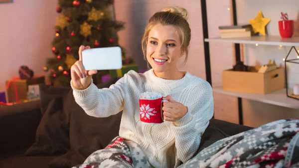 Joven mujer rubia sonriente cubierta de manta sentada en el sofá, sosteniendo una taza de cacao caliente con malvaviscos y tomando selfie en el teléfono inteligente en Navidad - foto de stock