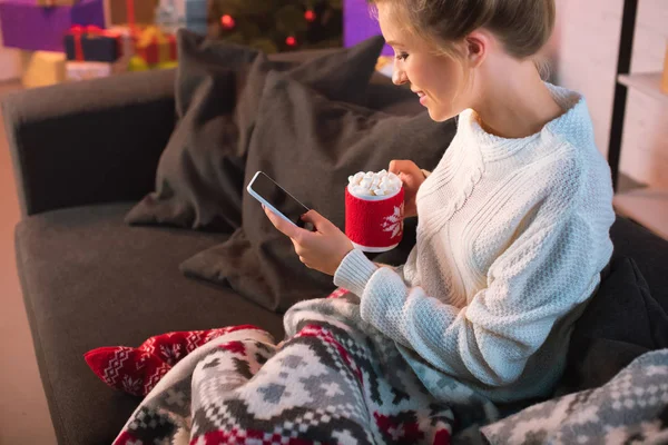 Jeune femme blonde tenant une tasse de chocolat chaud avec des guimauves et en utilisant un smartphone à la période de Noël — Photo de stock