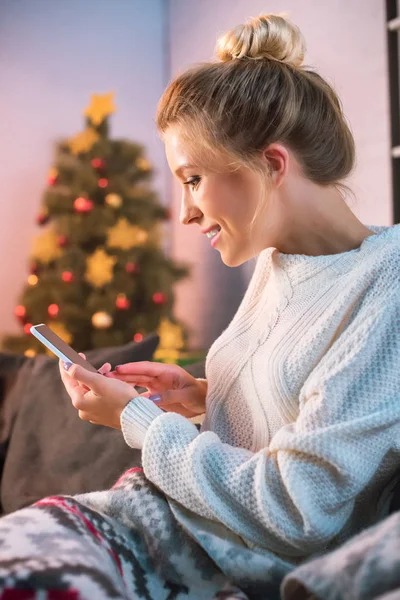 Счастливая молодая блондинка, сидящая на диване и пользующаяся смартфоном в это время — стоковое фото