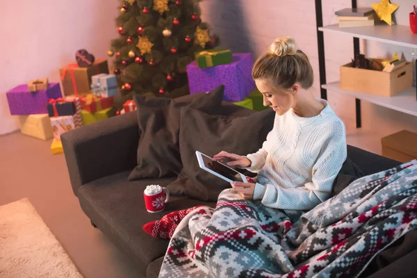 Sorrindo jovem loira sentada no sofá e usando tablet no Natal — Fotografia de Stock