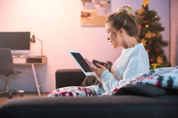 Вид збоку молода блондинка сидить на дивані і використовує планшет в різдвяний час — стокове фото