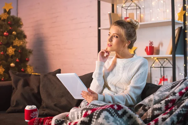 Pensive young blonde woman sitting on couch and using tablet at christmas time — Stock Photo