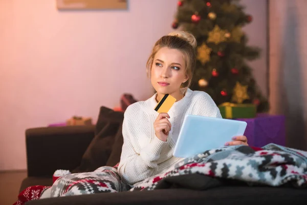 Chère jeune femme blonde assise sur un canapé tenant une carte de crédit et utilisant une tablette numérique pour faire du shopping en ligne à Noël — Photo de stock