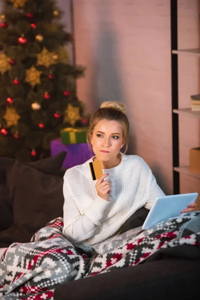 Pensive young blonde woman sitting on couch holding credit card and using digital tablet for online shopping at christmas time — Stock Photo