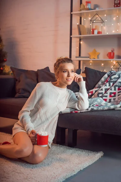 Hermosa mujer sentada en calcetines con dibujos navideños en la alfombra con taza de cacao - foto de stock