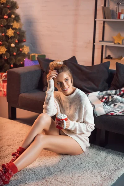 Mujer atractiva alegre sentada en la alfombra con taza de cacao caliente - foto de stock