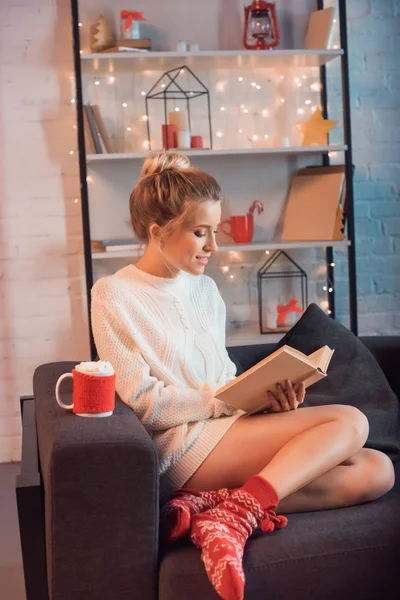Young blonde woman with cup of hot cocoa and marshmallows sitting on couch and reading book at christmas time — Stock Photo