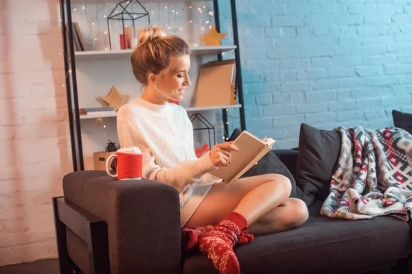 Smiling blonde young woman sitting on couch and reading book at christmas time — Stock Photo