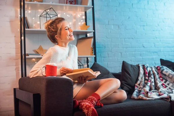 Riéndose joven rubia con taza de chocolate caliente y malvaviscos sentados en el sofá y leyendo el libro en Navidad - foto de stock