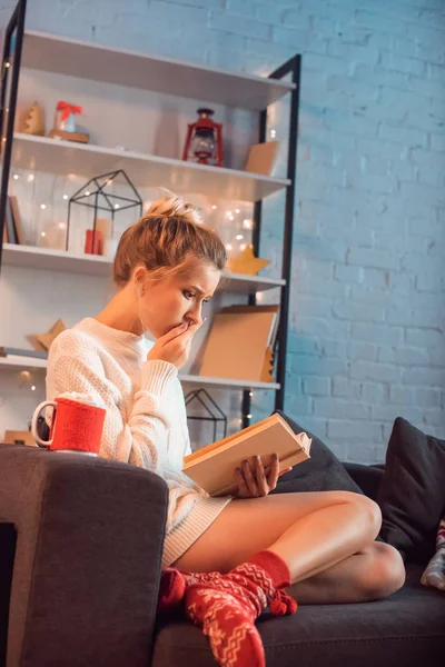 Mujer rubia joven asustada con una taza de cacao caliente y malvaviscos sentados en el sofá y leyendo el libro en Navidad - foto de stock