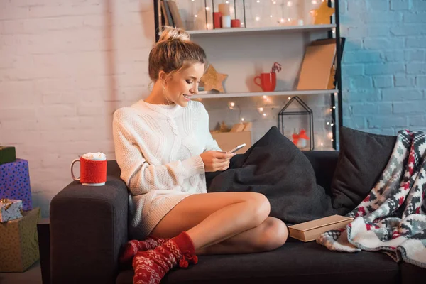 Sonriente joven rubia sentada en el sofá y usando el teléfono inteligente en Navidad - foto de stock