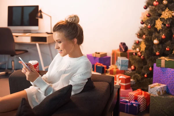 Smiling young blonde woman sitting on couch and using smartphone at christmas time — Stock Photo