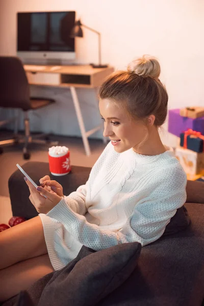 Smiling young blonde woman sitting on couch and using smartphone at christmas time — Stock Photo