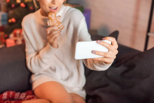 Cropped view of young woman sitting on couch, posing with gingerbread cookie and taking selfie on smartphone at christmas time — Stock Photo