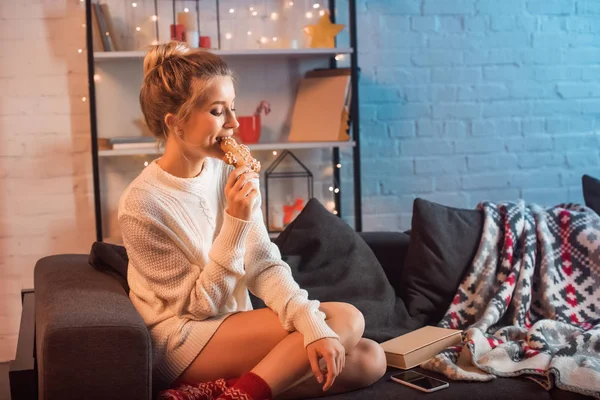 Beautiful young blonde woman sitting on couch and eating gingerbread cookie at christmas time — Stock Photo