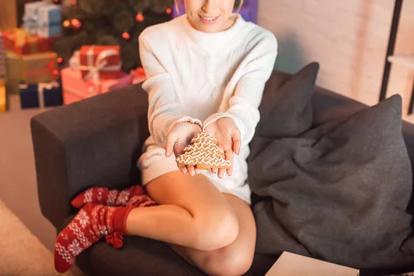 Vista ritagliata di sorridere giovane donna seduta sul divano e tenendo biscotti di pan di zenzero in mani outstreched al momento di Natale — Foto stock