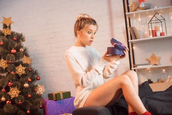 Surprised young blonde woman sitting on couch and opening present at christmas time — Stock Photo