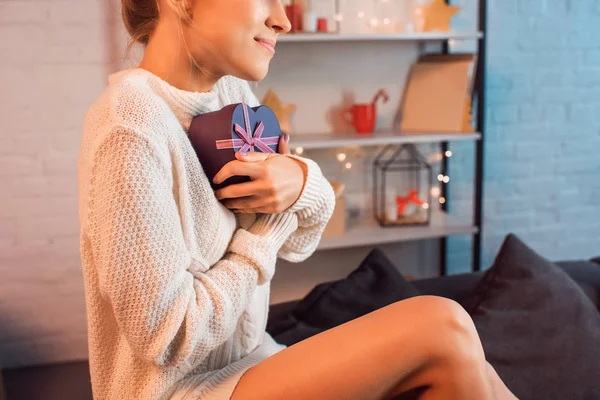 Vista recortada de la joven sonriente sentada en el sofá y sosteniendo presente en las manos en tiempo de Navidad - foto de stock