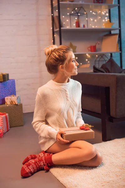 Blonde jeune femme assise, regardant loin et tenant présent à Noël — Photo de stock