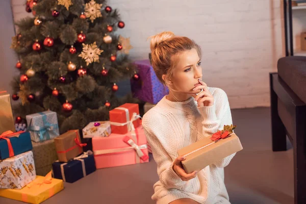 Mujer joven rubia pensativa sentada y sosteniendo presente en tiempo de Navidad - foto de stock