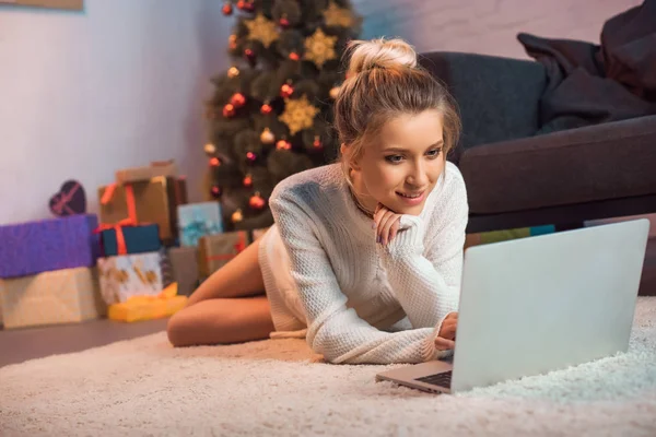 Усміхнена молода блондинка лежить на підлозі, використовуючи ноутбук в різдвяний час — стокове фото