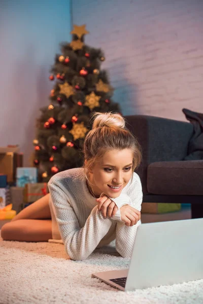 Feliz joven rubia tumbada en el suelo usando el ordenador portátil en Navidad - foto de stock