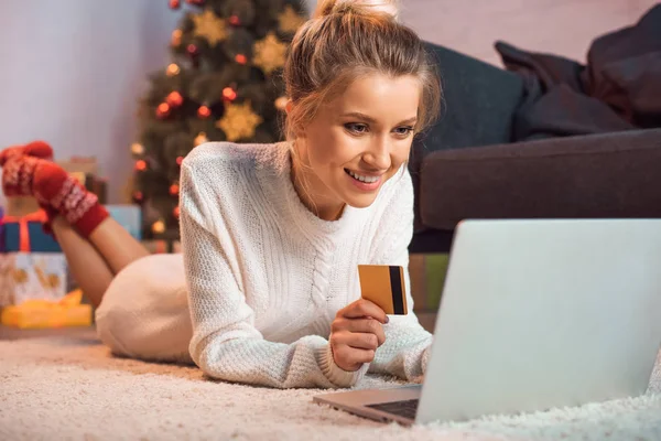 Sonriente joven rubia tumbada en el suelo, sosteniendo la tarjeta de crédito, usando el ordenador portátil y haciendo compras en línea en Navidad - foto de stock