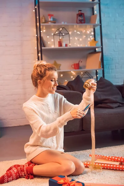 Smiling young blonde woman cutting ribbon with scissors for wrapping presents at christmas time — Stock Photo