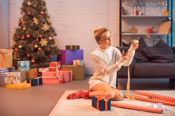 Улыбающаяся молодая блондинка перерезает ленточку ножницами для обертывания подарков на Рождество — Stock Photo