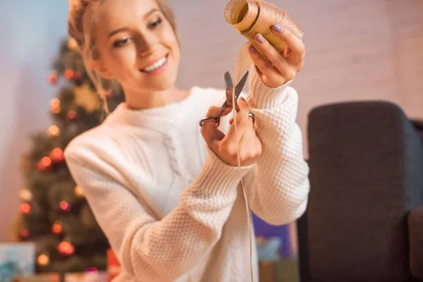 Selective focus of attractive young blonde woman cutting ribbon with scissors for wrapping presents at christmas time — Stock Photo