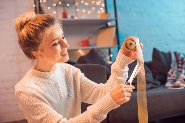 Beautiful smiling young blonde woman cutting ribbon with scissors for wrapping presents at christmas time — Stock Photo