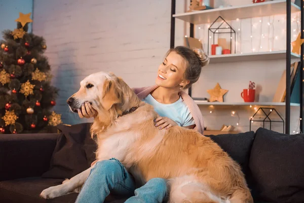 Hermosa feliz joven rubia sentada en el sofá y abrazando perro golden retriever en Navidad - foto de stock