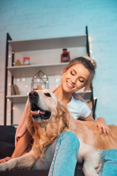 Hermosa alegre joven rubia sentada en el sofá y abrazando perro golden retriever en Navidad - foto de stock