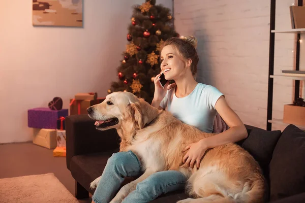 Belle jeune femme blonde assise sur le canapé avec chien golden retriever et parlant sur smartphone à Noël — Photo de stock