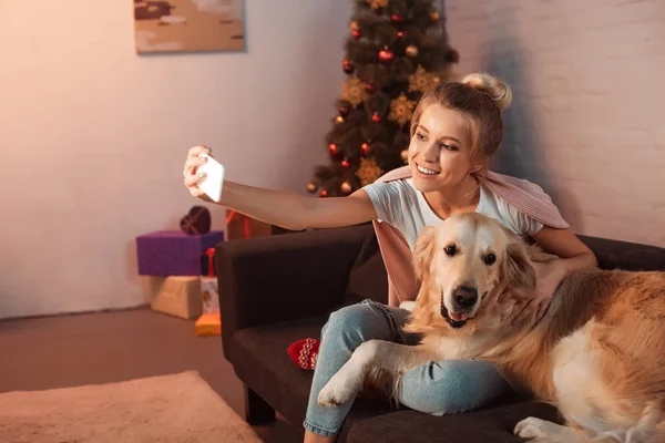 Belle jeune femme blonde assise sur le canapé avec chien golden retriever et prenant selfie sur smartphone à Noël — Photo de stock