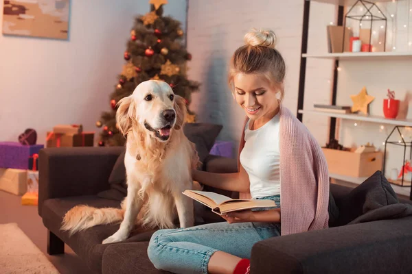 Hermosa joven rubia sentada en el sofá con perro golden retriever y libro de lectura en Navidad - foto de stock