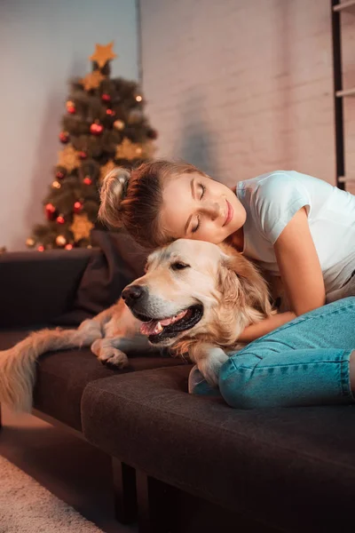 Hermosa sonriente joven rubia en el sofá abrazando perro golden retriever en Navidad - foto de stock