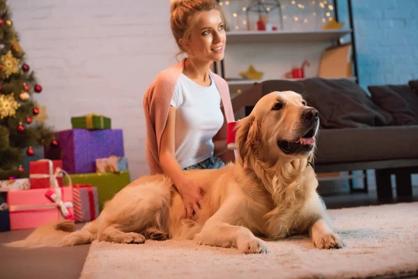 Hermosa sonriente joven rubia sentada en el suelo con perro golden retriever en Navidad - foto de stock