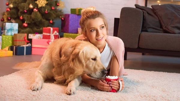 Beautiful young blonde woman lying on floor with golden retriever dog and cup of hot cocoa with marshmallows at christmas time — Stock Photo
