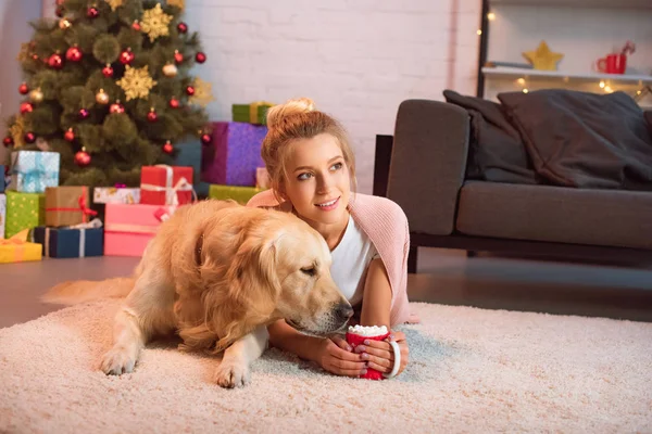 Belle jeune femme blonde couchée sur le sol avec chien récupérateur d'or et tasse de cacao chaud avec guimauves à Noël — Photo de stock