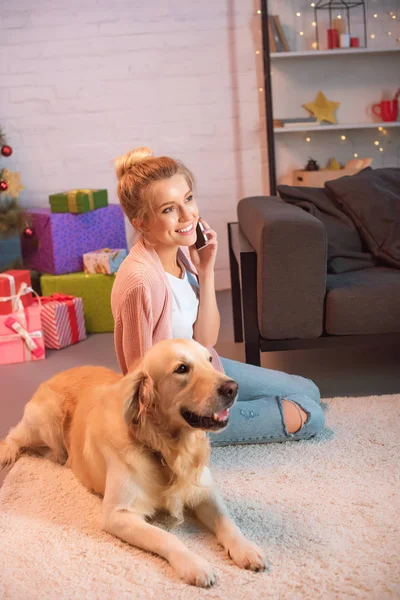 Beautiful young blonde woman sitting on floor with golden retriever dog and talking on smartphone at christmas time — Stock Photo