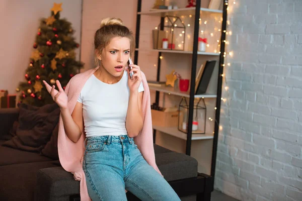 Сердитая молодая блондинка, сидящая на диване и разговаривающая по смартфону на Рождество — стоковое фото