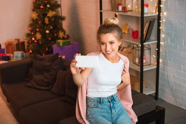 Smiling young blonde woman sitting on couch and taking selfie on smartphone at christmas time — Stock Photo