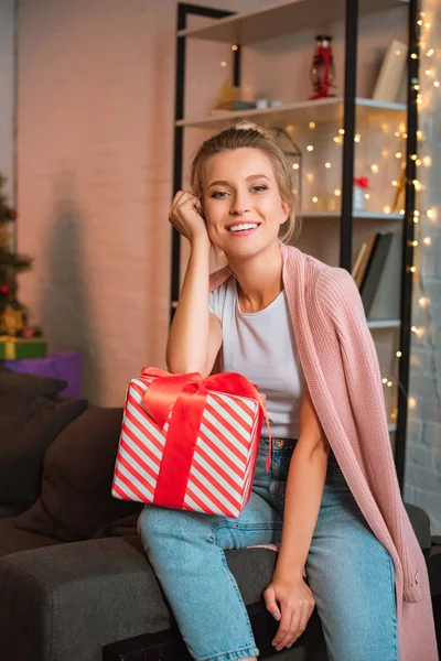 Happy young blonde woman sitting on couch with present and looking at camera at christmas time — Stock Photo