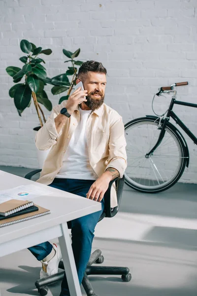 Hombre de negocios barbudo sentado en la silla de oficina y hablando en el teléfono inteligente - foto de stock